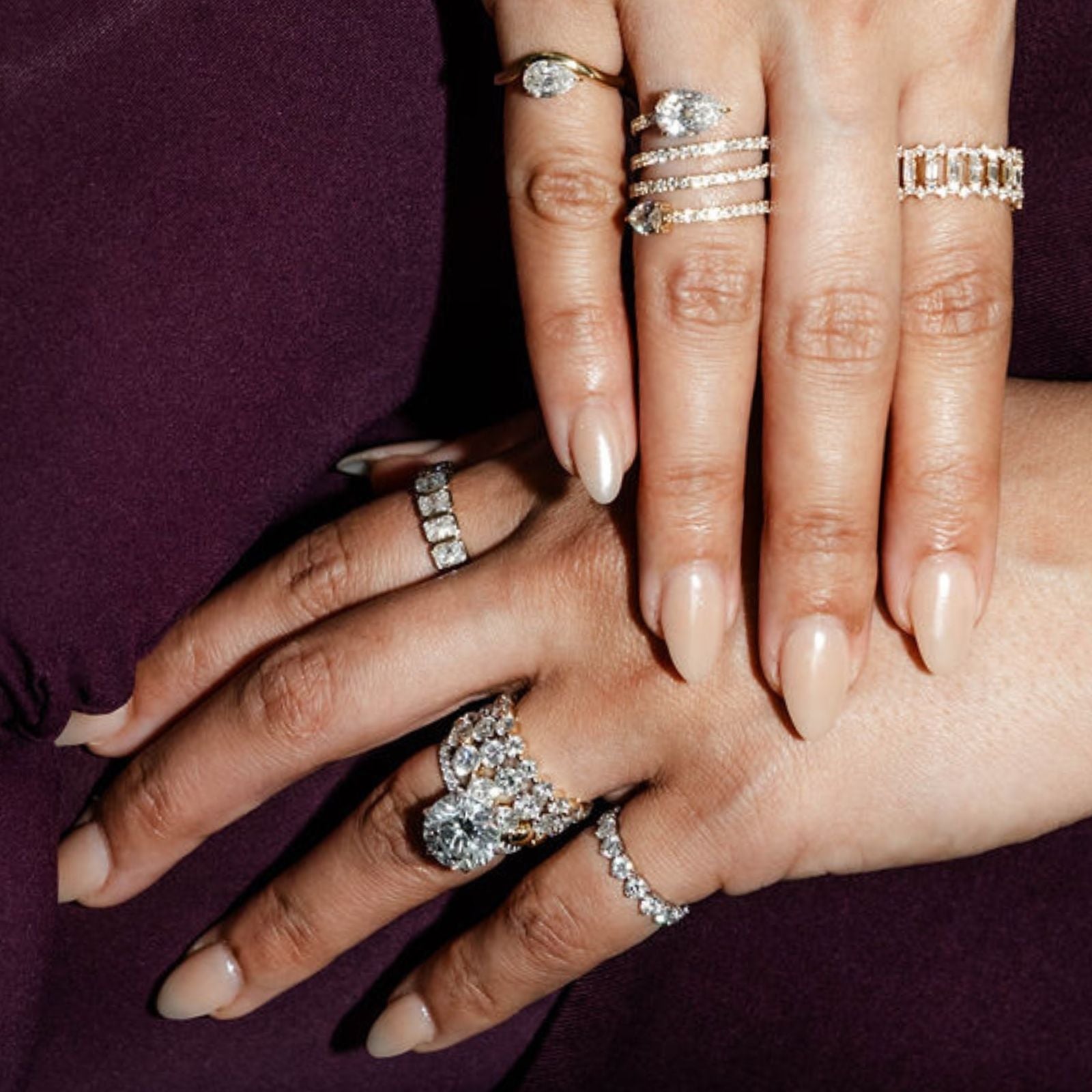 Two hands layered together wearing large diamond rings and eternity bands against a dark background.