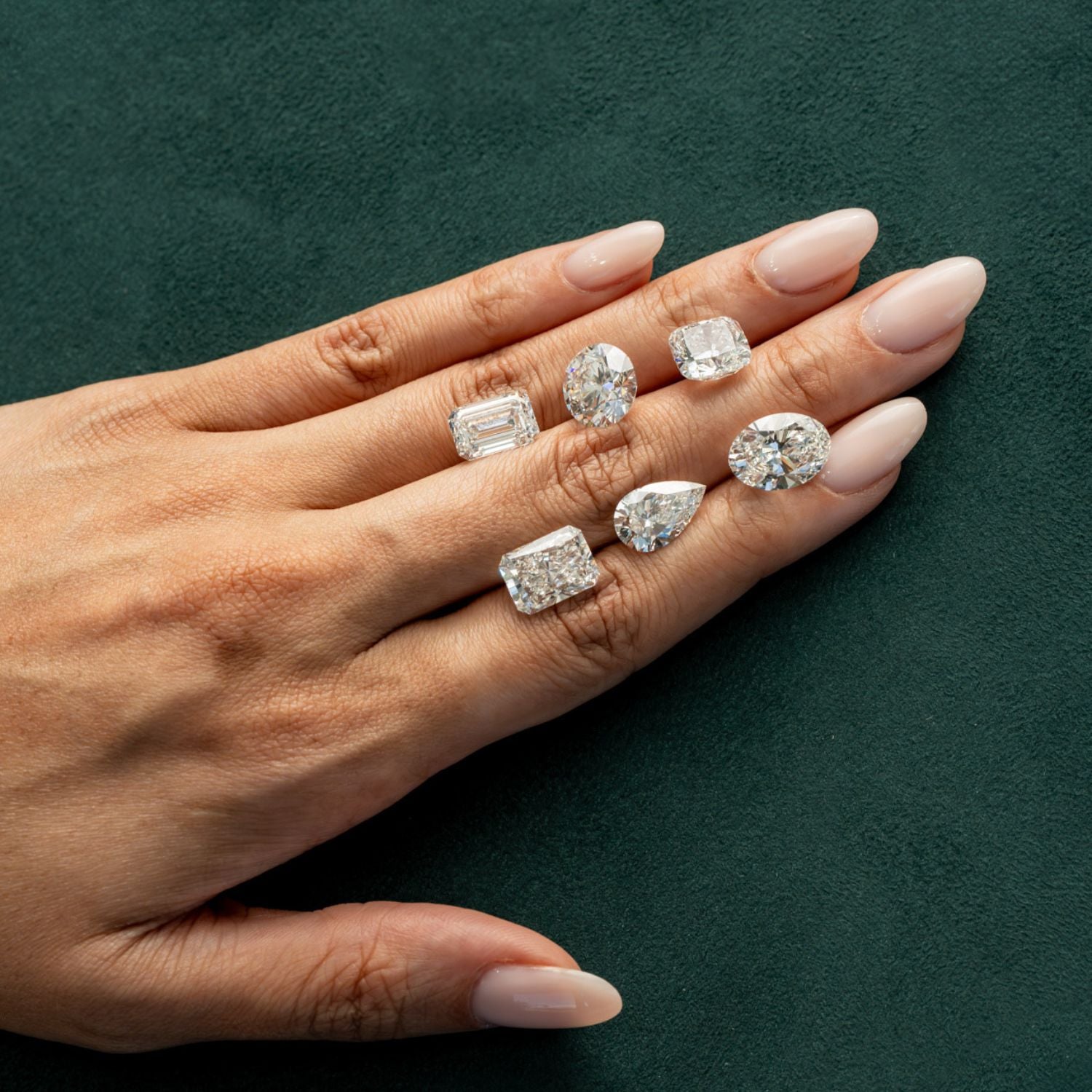 Hand displaying six loose diamonds in different cuts including emerald, oval, cushion and pear shapes on a green velvet surface.