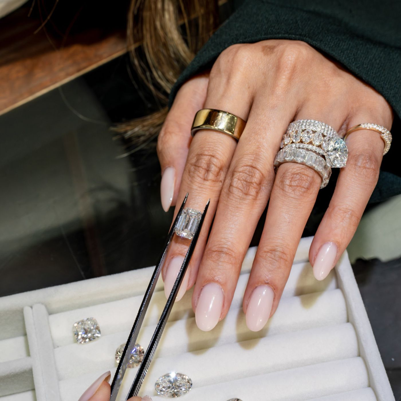 Jeweler presenting loose diamonds on a tray using tweezers, with stacked diamond rings visible on hand.
