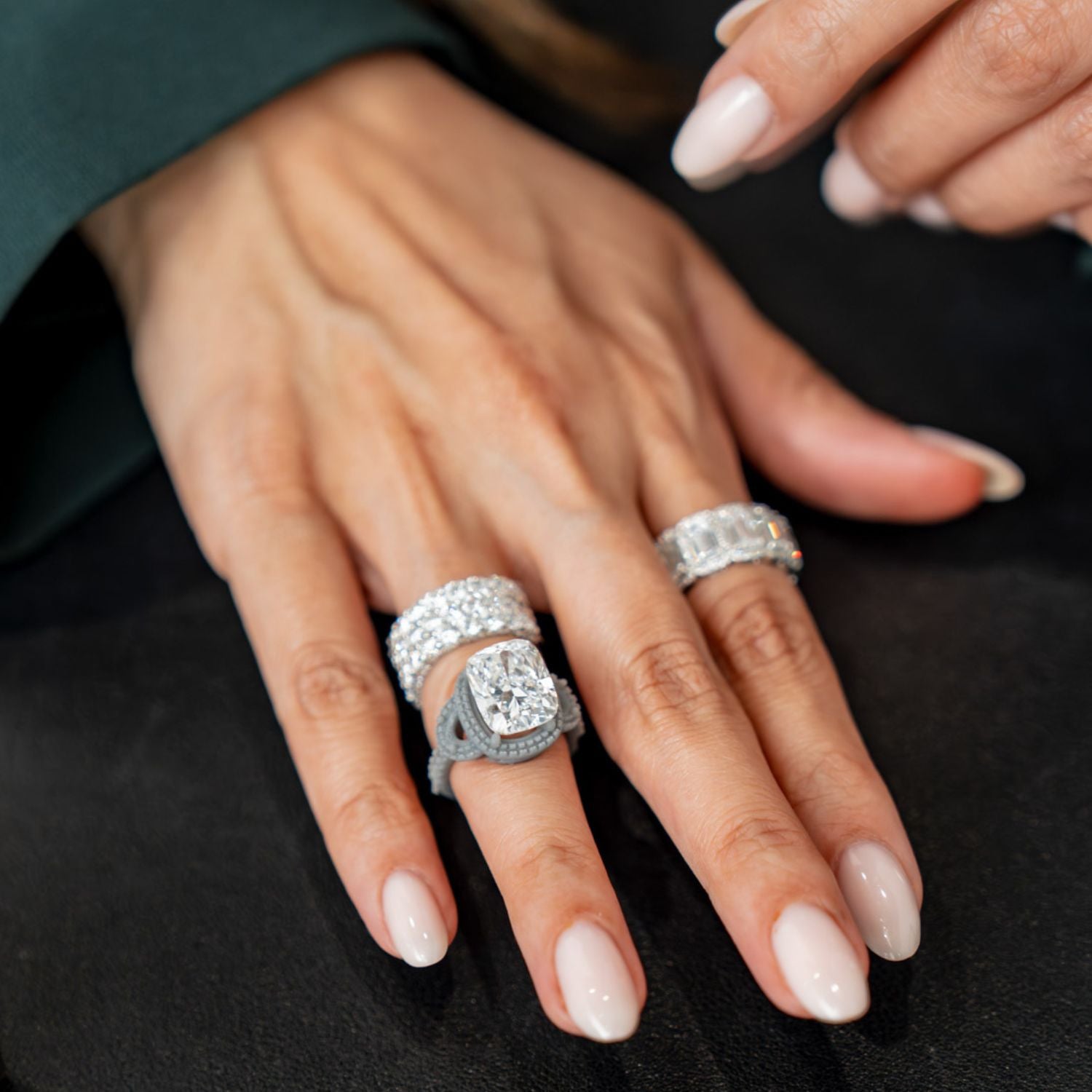 Hand wearing large cushion-cut and emerald-cut diamond rings displayed against a dark velvet surface.