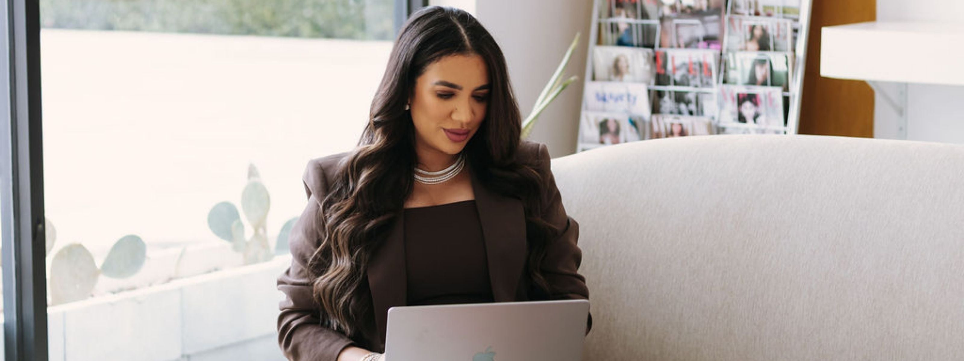 Founder in a brown blazer and diamond necklace working on a laptop in a bright modern lounge setting.